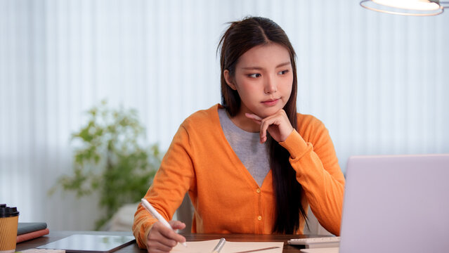 Young Asian student writing notes in notebook while studying at home desk with laptop and coffee