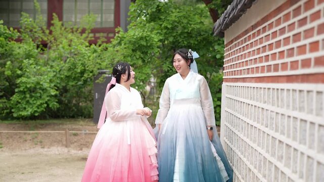 Two women in traditional Korean dresses laugh together. They walk beside a stone wall near greenery. Their joyful expressions suggest a pleasant, cultural outing. Ideal for travel, heritage