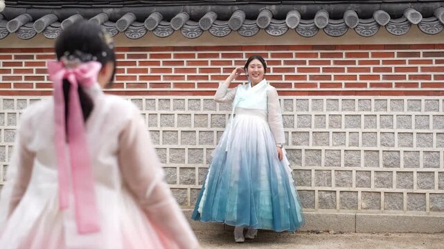 Two women in traditional Korean dresses pose gracefully. One in soft pink, the other in blue gradient. They stand before a classic tiled brick wall. Their joyful expressions show cultural pride