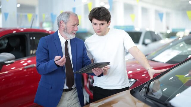Elderly car salesman discusses terms and demonstrates car to young male buyer at car dealership