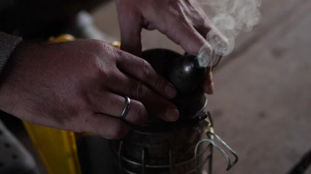 A close-up cinematic shot of a beekeeper using a gas torch to light charcoal inside a traditional bee smoker tool at an apiary.