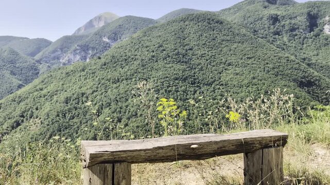 Panchina rustica in legno posizionata su un prato verde con vista su colline e montagne ricoperte di vegetazione sotto un cielo limpido