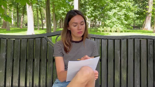 A young girl thoughtfully solves a test while sitting on a bench in a park.