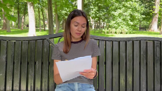 Young woman sitting on a park bench reading a document and writing notes with a pencil. Reviewing important notes in a peaceful outdoor setting.
