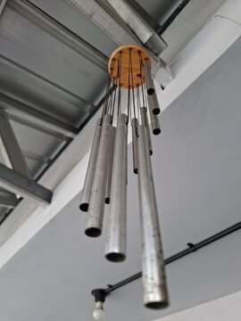 bottom-up perspective of a metallic wind chime made of several silver aluminum tubes hanging from a round wooden base underneath a corrugated ceiling.