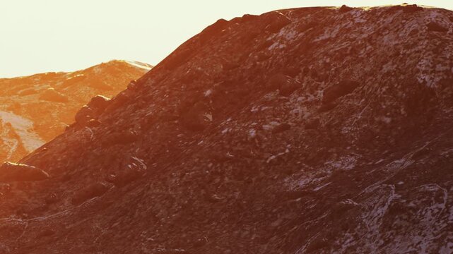 The sun sets over rugged mountains creating shadows on the rocky surface. The landscape shows a mixture of light and dark areas, highlighting the natural features of the terrain.
