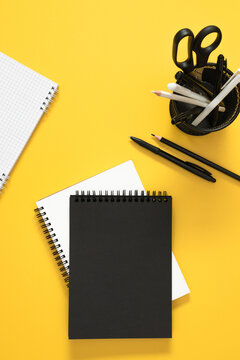 Vertical Flat Lay with Blank Black Cover Notebook, Grid Notepad, and Stationery Organizer on Bright Yellow Background. Contrast Minimalist Office and Back to School Concept. Top View.