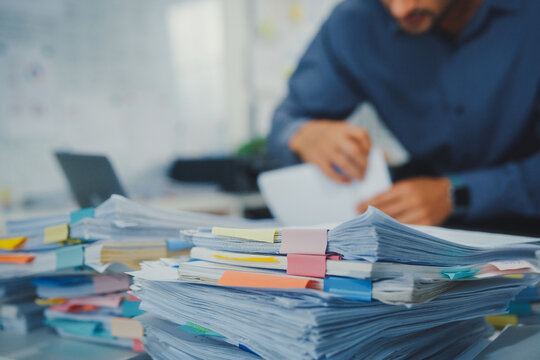 Closeup overworked young Latin businessman reading document in large stack of paperwork file at office desk. Tax audit deadline, accounting stress, corporate burnout, bookkeeping, bureaucracy concept.