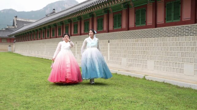 Two women in colorful hanboks walk gracefully on grass. They smile softly, radiating calm joy and cultural pride. Traditional buildings loom behind them, framed by mountains