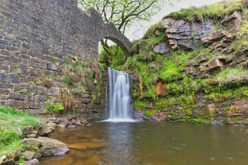 A scenic waterfall cascades into a pool, flowing under a stone arch bridge, framed by an old stone wall and lush green moss - Cheesden Lumb Mill, Lancashire, UK © Robert