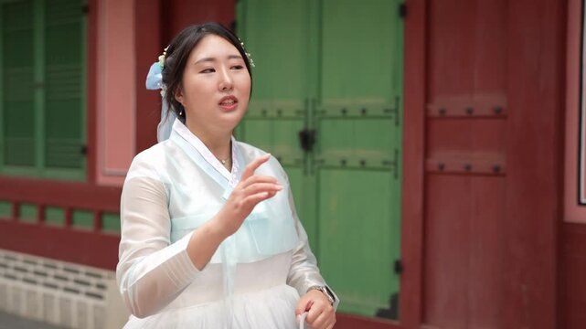 Woman in traditional Korean attire speaks. She gestures with hands while standing outside. Red and green gates form background. Her expression shows engagement and passion