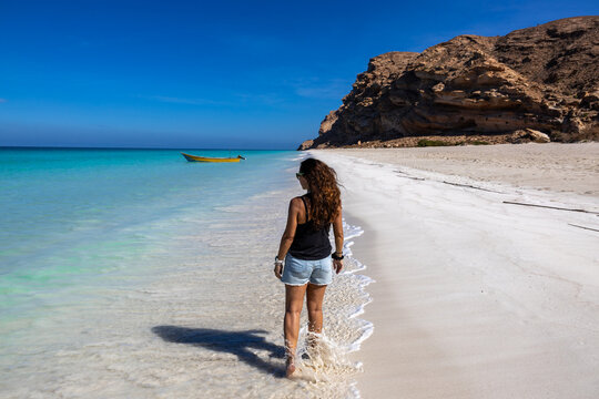 A woman walking barefoot along the white sandy shoreline of Shuab Beach on Socotra Island, Yemen, with turquoise waters and dramatic cliffs in the background.