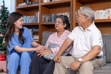 Naklejka premium Happy caregiver woman supporting senior couple sitting on sofa in cozy living room with bookcase smiling feeling comforted and relaxed during home visit healthcare consultation service