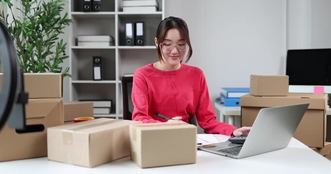 Young asian woman entrepreneur organizing customer orders at a home office, surrounded by stacks of cardboard boxes, processing online sales and managing shipping logistics for her small business