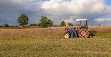 Obraz premium Farmer in workwear inspecting and repairing an old red tractor in a rural field, illustrating agricultural maintenance, mechanical problems and authentic countryside work