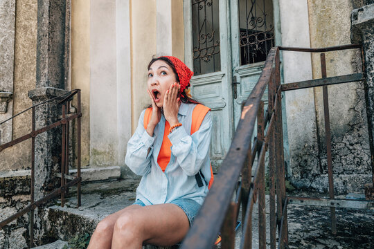 Young backpacker woman sitting on old stairs, showing surprised and shocked expression. Exploring an abandoned building during travel