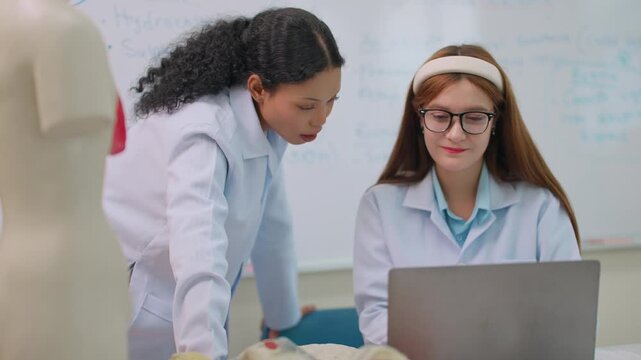 Two students in lab coats collaborate near a laptop. One student leans in, guiding the other's attention. Anatomical models stand beside them, near a whiteboard. Both students look engaged