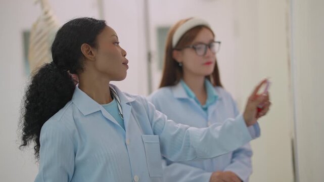 Young woman writes on whiteboard in classroom. Another student watches her intently. Scene suggests active learning and discussion. Footage captures educational moment, focused and engaged