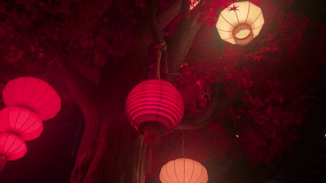 Red and white paper lanterns hanging from tree in traditional Chinese garden. Cultural decoration for Lunar New Year and Asian festival celebration. Glowing night scene in town.