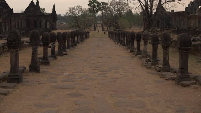 Camera pushes forward down the stone walkway at Vat Phou, passing rows of boundary posts between palace ruins and dry trees.