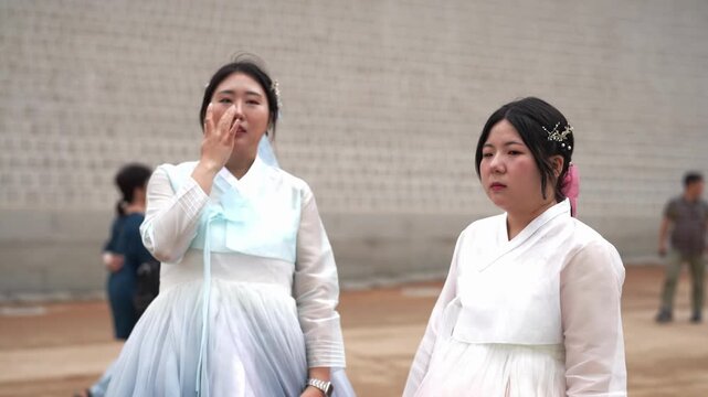Two women in traditional Korean attire laugh together. One covers her mouth, the other claps her hands. They stand before a simple brick wall. Their joy radiates in casual, joyful interaction