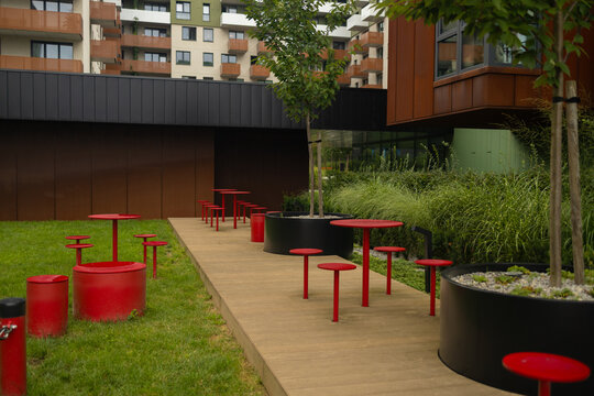 A contemporary apartment complex inner courtyard featuring a stylish wooden deck pathway, bright red tables with chairs, and modern landscaping with green grass.