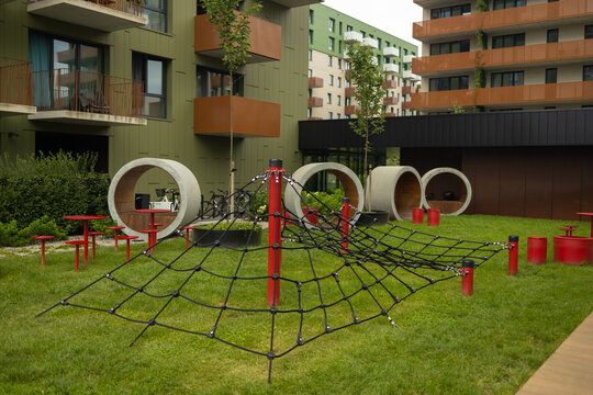 A contemporary outdoor kids playground featuring a black rope climbing net and concrete play tunnels on a green lawn in a modern residential apartment building courtyard.