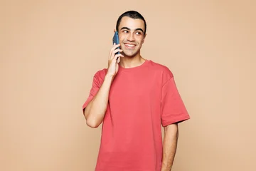 Young middle eastern man he wears red t-shirt casual clothes talk speak on mobile cell phone conducting pleasant conversation isolated on plain pastel light beige background studio. Lifestyle concept. © ViDi Studio