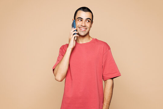 Young middle eastern man he wears red t-shirt casual clothes talk speak on mobile cell phone conducting pleasant conversation isolated on plain pastel light beige background studio. Lifestyle concept.
