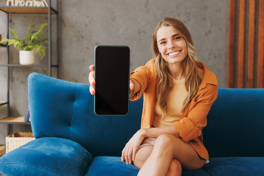 Young happy woman wear brown shirt hold in hand use mobile cell phone blank screen workspace sit on blue sofa stay at home hotel flat rest relax spend free spare time in living room indoors grey wall