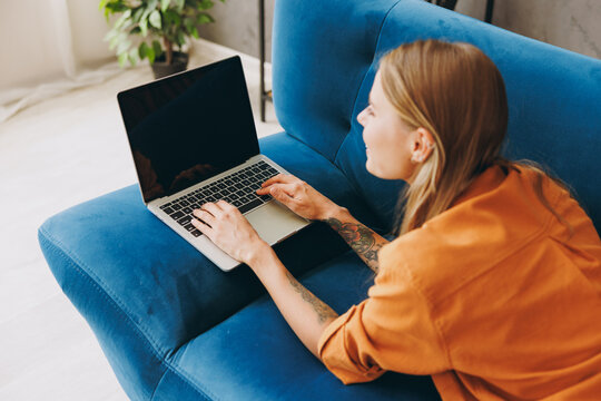Back view young IT woman in brown shirt hold use work laptop pc computer blank screen workspace area lay down blue sofa couch stay at home flat rest relax spend free spare time in living room indoor