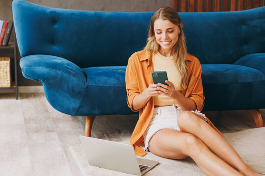 Happy young woman wear brown shirt use work laptop pc computer hold mobile phone sit at blue sofa stay at home hotel flat rest relax spend free spare time in living room indoors People lounge concept