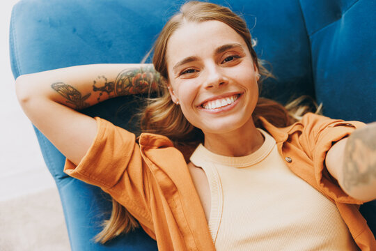Close up young woman wear brown shirt do selfie shot pov on mobile cell phone lay down blue sofa stay at home hotel flat rest relax spend free spare time in living room indoors. People lounge concept.