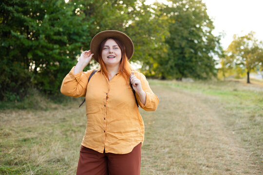 A cheerful young female tourist wearing a brown hat and an orange shirt adjusts her backpack while hiking through a lush green forest on a sunny day.