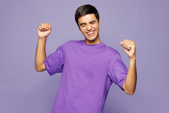 Young Caucasian man he wears violet t-shirt casual clothes doing winner gesture celebrate clenching fists dancing isolated on plain pastel light purple background studio portrait. Lifestyle concept.