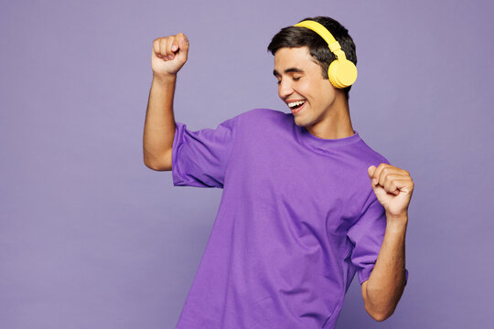 Young happy Caucasian man he wear violet t-shirt casual clothes listen to music in headphones raise up hands dance isolated on plain pastel light purple background studio portrait. Lifestyle concept.