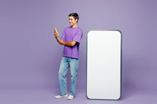 Full body young happy man he wear violet t-shirt casual clothes big huge blank screen mobile cell phone with workspace copy space mockup area use smartphone isolated on plain light purple background.