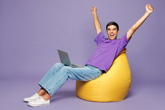 Full body young IT man he wear violet t-shirt casual clothes sit in bag chair hold use work on laptop pc computer do winner gesture isolated on plain pastel light purple background. Lifestyle concept.