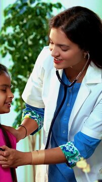 Indian Asian playful female doctor or pediatrician engaging young girl patient by letting her hear own heartbeat using stethoscope in a warm and friendly clinic atmosphere