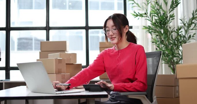 Young asian woman entrepreneur organizing customer orders at a home office, surrounded by stacks of cardboard boxes, processing online sales and managing shipping logistics for her small business