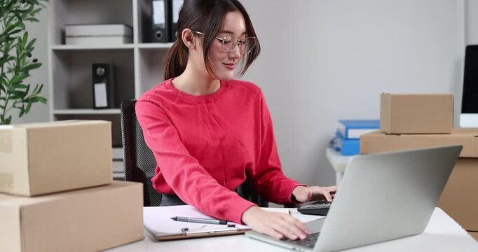 Young asian woman entrepreneur organizing customer orders at a home office, surrounded by stacks of cardboard boxes, processing online sales and managing shipping logistics for her small business