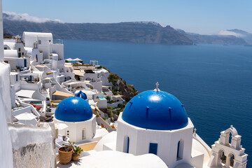 Famous white churches with blue domes and tiered bell tower in Oia, Santorini. Traditional Cycladic buildings cascade down volcanic cliffs overlooking the blue Aegean Sea on a sunny day. © Pasha