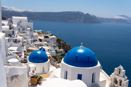 Famous white churches with blue domes and tiered bell tower in Oia, Santorini. Traditional Cycladic buildings cascade down volcanic cliffs overlooking the blue Aegean Sea on a sunny day.