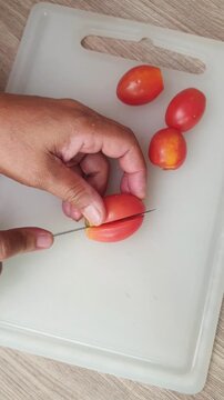 A person using a kitchen knife to slice fresh red tomatoes on a white cutting board for meal preparation