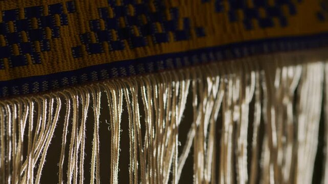 Decorative woven band in mustard yellow and deep blue with hanging off-white fringe threads, side lit for rich texture, pattern contrast, and tactile handmade detail