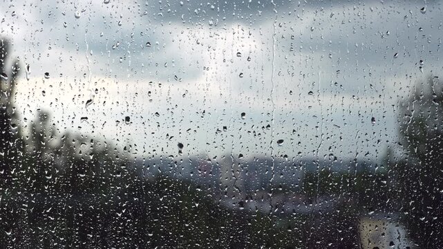 Close-up video of raindrops slowly running down a wet window during rainy weather. Water droplets sliding across the glass create a calm, moody atmosphere with a soft blurred background and natural da
