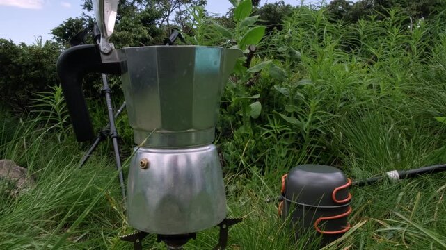 A horizontal close up shot of a classic moka pot brewing hot coffee on a portable stove in lush green grass, perfect for travel storytelling, morning wellness, and lifestyle marketing.