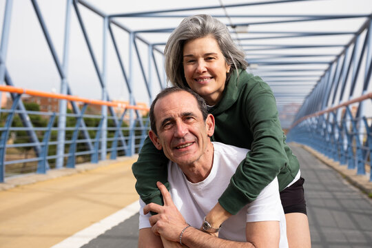 Happy mature couple piggybacking, bonding and smiling on an urban bridge