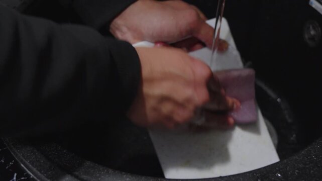 Person washing plastic cutting board with sponge under running water