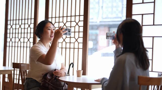 Two women smile, capturing moments with smartphones. Soft light filters through wooden lattice windows. Cafe setting suggests casual, friendly connection. Ideal for social, lifestyle